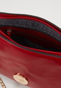 Borsa in finta pelle rossa con un accento dorato del logo e una tasca con zip. L'interno presenta una fodera scura con motivi sottili. Inclusa la tracolla a catena.