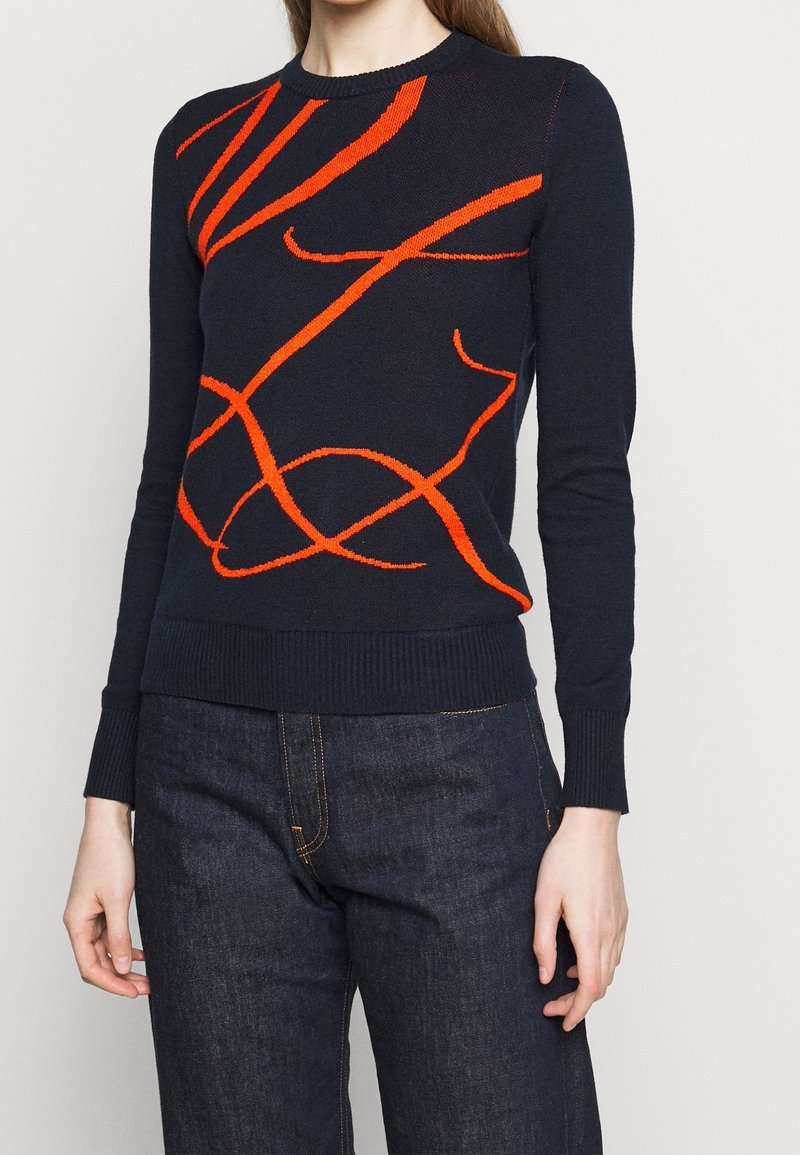Femme portant un pull noir ajusté avec des lignes orange abstraites, associée à un jean bleu foncé sur un fond uni.