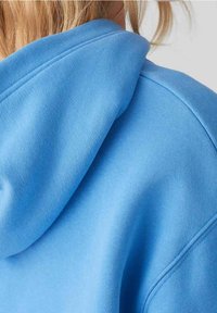 Osoba s blond vlasy, oblečená v světle modré mikině s kapucí, viditelně ukazující zadní část a oblast kapuce se zvýrazněnými švy.