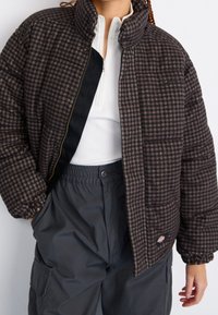 Doudoune à carreaux brun avec un col montant, fermeture éclair, poignets élastiques et poches avant, portée sur un haut blanc et un pantalon gris.