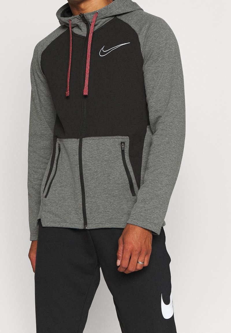 Homme portant un sweat à capuche zippé Nike noir et gris avec des cordons rouges et un pantalon noir arborant un logo Nike blanc sur la cuisse gauche.