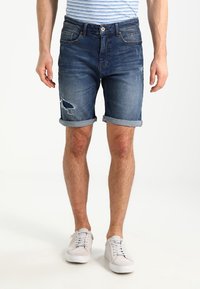 Man som bär uppvikta mörkblå slitna denimshorts och vita sneakers, stående mot en enkel ljus bakgrund.
