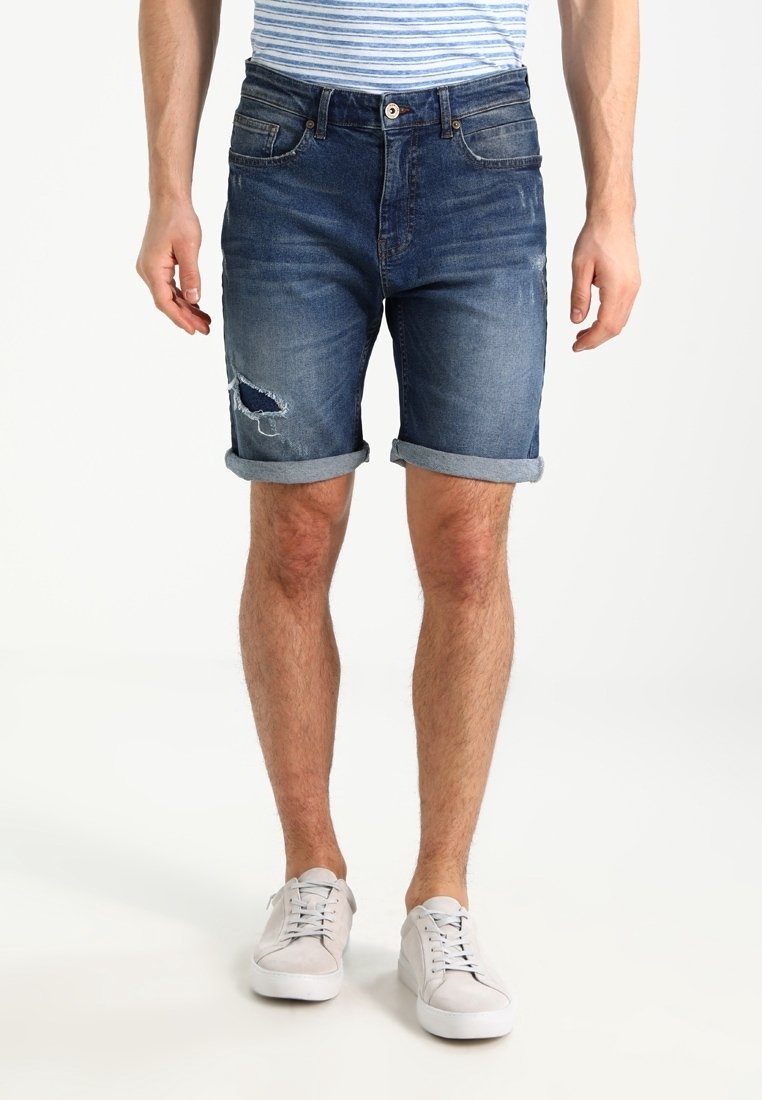 Man som bär uppvikta mörkblå slitna denimshorts och vita sneakers, stående mot en enkel ljus bakgrund.