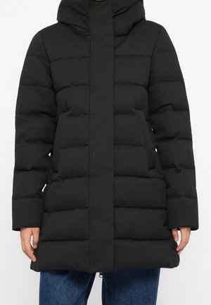 Veste matelassée noire avec col montant et fermeture avant dissimulée portée sur un jean bleu, montrant le torse et les bras.