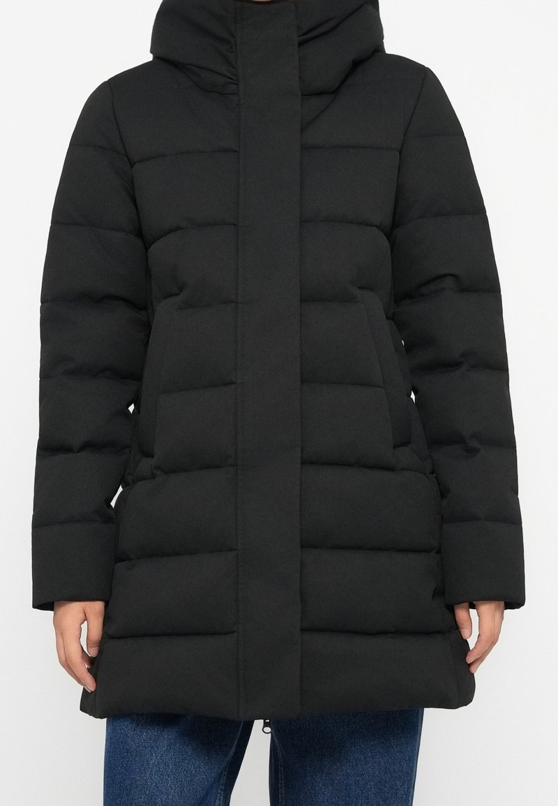 Veste matelassée noire avec col montant et fermeture avant dissimulée portée sur un jean bleu, montrant le torse et les bras.
