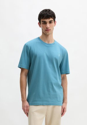 Giovane uomo in piedi che indossa una T-shirt blu a maniche corte semplice e pantaloni beige su sfondo bianco.