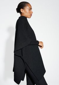 Zwarte gebreide poncho met een losse, asymmetrische vorm en geribbelde textuur. Voorzien van lange mouwen en een hoge hals, ontworpen voor een oversized pasvorm.