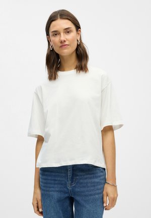 Femme aux cheveux bruns mi-longs portant des boucles d'oreilles argentées en anneau, un t-shirt blanc simple à manches courtes, un jean bleu et un bracelet argenté.