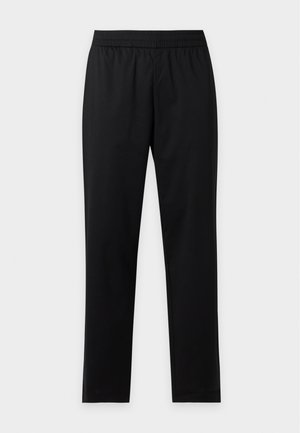 Homme portant un polo bleu clair à manches courtes, un pantalon noir et des chaussures noires, debout les mains dans les poches devant un fond uni.