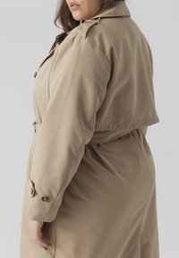 Béžový trench coat se strukturovaným límcem, zapínáním na knoflíky a páskem v pase. Vyrobený z hladké, lehké látky s jemným leskem.