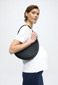 Czarna skórzana torebka w kształcie półksiężyca o fakturowanej powierzchni i regulowanym pasku, trzymana na tle białej bluzki. Czysty, nowoczesny design z minimalnymi detalami.