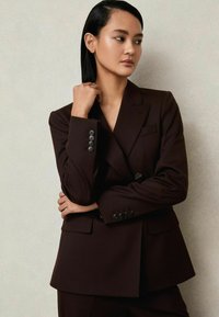 Blazer ajusté marron foncé avec un design croisé, des revers en pointe et six boutons. Présente des poches avant et une silhouette structurée.