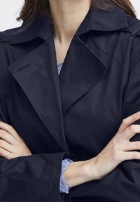 Námořnický trenčkot s vykrojeným límcem, vyrobený z lehkého materiálu, s texturovaným povrchem. Pod ním jsou viditelné modré krajkové manžety.