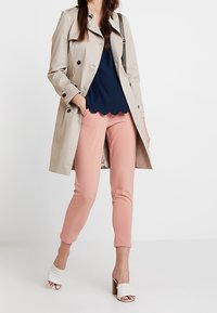 Gabardina beige con botones de doble botonadura, blusa azul marino con volantes, pantalones rosas ajustados y sandalias de tacón blancas tejidas.