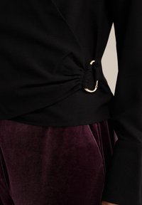 Top nero con design drappeggiato, caratterizzato da un accentuato anello dorato e un dettaglio di tessuto arricciato. Abbinato a pantaloni in velluto viola morbido.