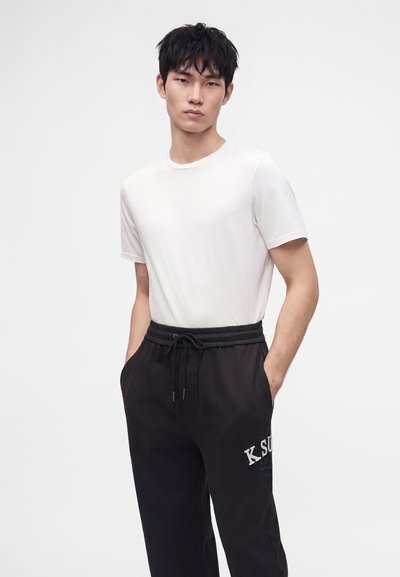 Νέος άνδρας με απλό λευκό t-shirt και μαύρη φόρμα, με τα χέρια στις τσέπες, μπροστά σε απλό φωτεινό φόντο.
