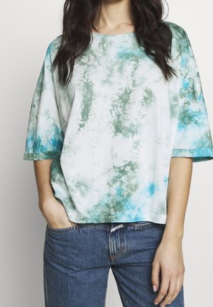 Kvinde iført en løs, kortærmet tie-dye skjorte med grønne og blå mønstre, kombineret med blå denimjeans, stående med den ene hånd i lommen.