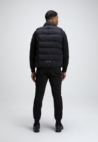 Czarna kamizelka puchowa z wysokim kołnierzem, pikowanym wzorem i logo z tyłu, noszona na dopasowanym czarnym dresie. Gładka tekstura i dopasowany krój.