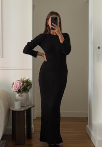 Črna rebrasta obleka z dolgimi rokavi in prilegajočim se krojem ter detajlom na robu. Oblečena jo ima oseba, ki drži telefon, v ozadju pa so rastlina in dekor.