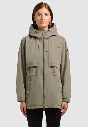 Kobieta w jasnej oliwkowozielonej kurtce z kapturem, z zamkiem błyskawicznym z przodu, kieszeniami z klapkami i elastycznymi mankietami, stojąca na jednolitym tle.