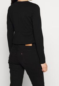 Personne portant un haut noir ajusté à manches longues et un jean noir taille haute, vue de dos sur un fond clair uni.