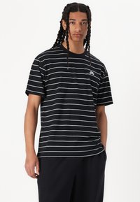 Schwarz-weißer gestreifter T-Shirt aus Baumwolle; hat einen Rundhalsausschnitt und kurzärmelige Ärmel. Kleines Logo auf der Brust. Modell trägt dunkle Hosen.