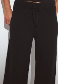 Moteris dėvi juodas plačias kelnes su raišteliu juosmenyje, stovėdama prieš paprastą baltą foną.