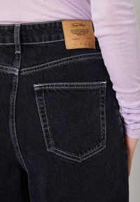 Tuva no tumši zilām džinsa biksēm ar redzamu šuvi un brūnu ādas zīmola etiķeti, valkātas ar gaiši violetu garroku krekliņu.