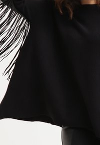 Kvinna klädd i en svart texturerad poncho med lång fransdetalj på ena ärmen och svarta tajta byxor.