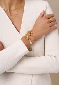 Blazer tricotato bianco con texture e bottone dorato, collane dorate a strati e bracciali dorati, inclusi un bracciale a catena con chiusura a linguetta.
