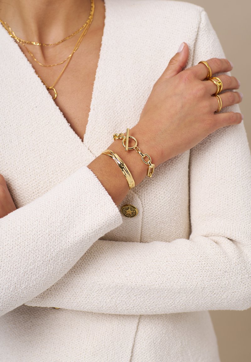 Blazer tricotato bianco con texture e bottone dorato, collane dorate a strati e bracciali dorati, inclusi un bracciale a catena con chiusura a linguetta.