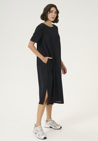 Schwarz lockeres Kleid mit kurzen Ärmeln, seitlichen Taschen und Schlitzen am Saum. Getragen mit grauen und weißen Sportschuhen.
