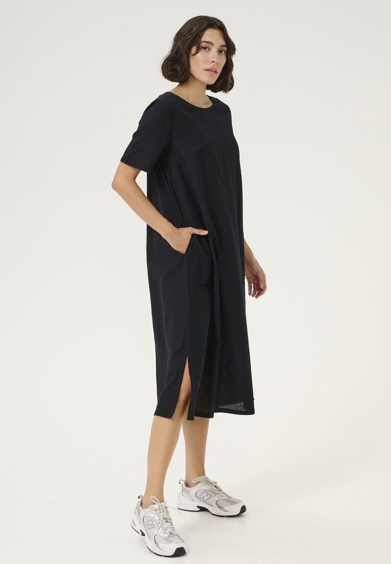 Schwarz lockeres Kleid mit kurzen Ärmeln, seitlichen Taschen und Schlitzen am Saum. Getragen mit grauen und weißen Sportschuhen.
