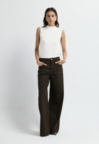 Top bianco senza maniche con collo alto, abbinato a pantaloni a gamba larga marrone scuro con decorazioni di perline metalliche e tasche frontali.
