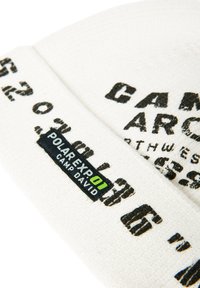 White knit beanie with black text pattern and a black label featuring green and white branding. Text is unevenly printed for a worn effect.