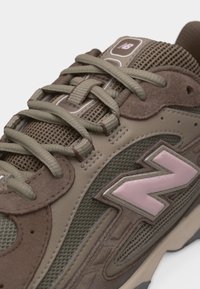 Καφέ και μπεζ αθλητικό παπούτσι New Balance με πάνελ από δίχτυ και σουέντ, μπεζ κορδόνια και ροζ λογότυπο "N" στο πλάι.
