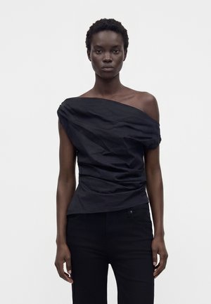 Femme aux cheveux courts et bouclés portant un haut noir asymétrique épaules découvertes et un pantalon noir, debout devant un fond clair uni.