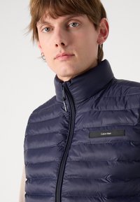 Jūras spēku zila mīksti pildīta veste ar horizontālu quilted dizainu, augstu apkakli un zīmola etiķeti. Iekļauj priekšējo rāvējslēdzēju un gludu, spīdīgu tekstūru.