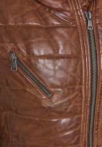 Chaqueta de cuero marrón con textura acolchada, que presenta un bolsillo con cremallera lateral y un cierre central con cremallera, mostrando costuras y detalles duraderos.
