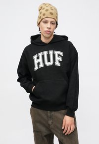 Μαύρο φούτερ με κουκούλα και λευκό λογότυπο "HUF", με τσέπη καγκουρό και ριμπ μανσέτες. Συνδυάζεται με καφέ υφασμάτινο cargo παντελόνι και σκουφάκι με πουά.