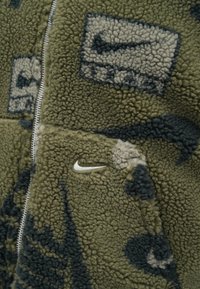 Zelena i crna uzorčasta flis jakna sa srebrnim patentnim zatvaračem i bijelim Nike swoosh logotipom izvezenim na džepu s prednje strane.