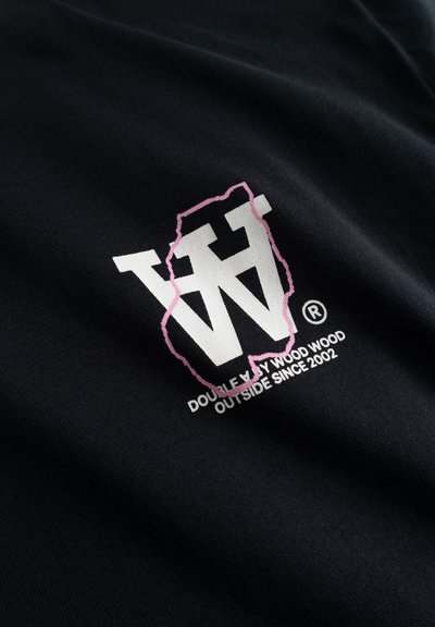 Logo blanc double W avec contour rose et texte "Double By Wood Wood Outside Since 2002" sur tissu noir.