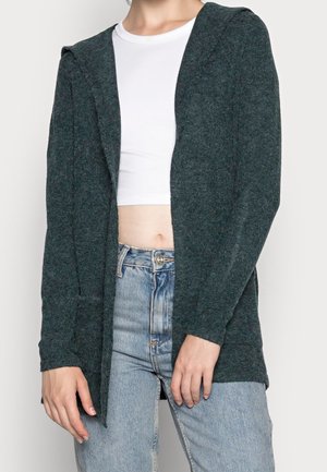 Vrouw draagt een donkergroene gebreide open cardigan over een wit cropped top en lichtblauwe hoge taille jeans tegen een witte achtergrond.