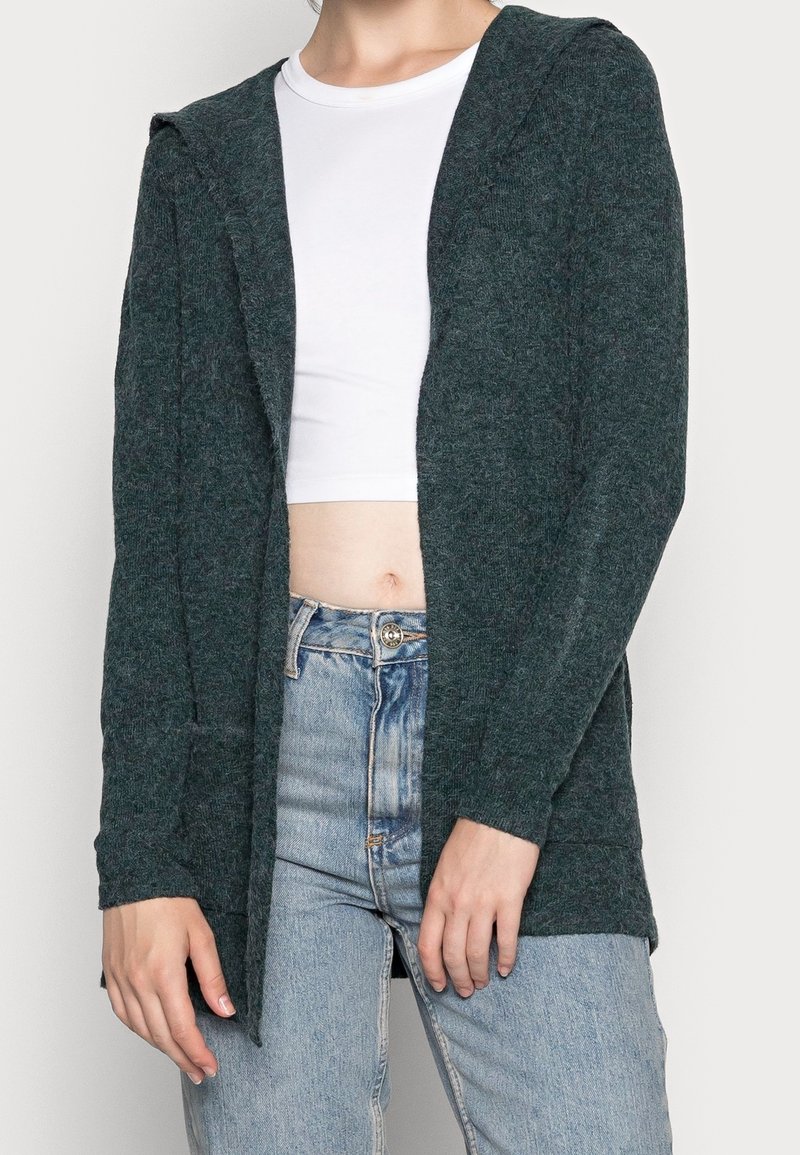 Femme portant un cardigan en maille vert foncé ouvert sur un crop top blanc et un jean taille haute bleu clair sur fond blanc.