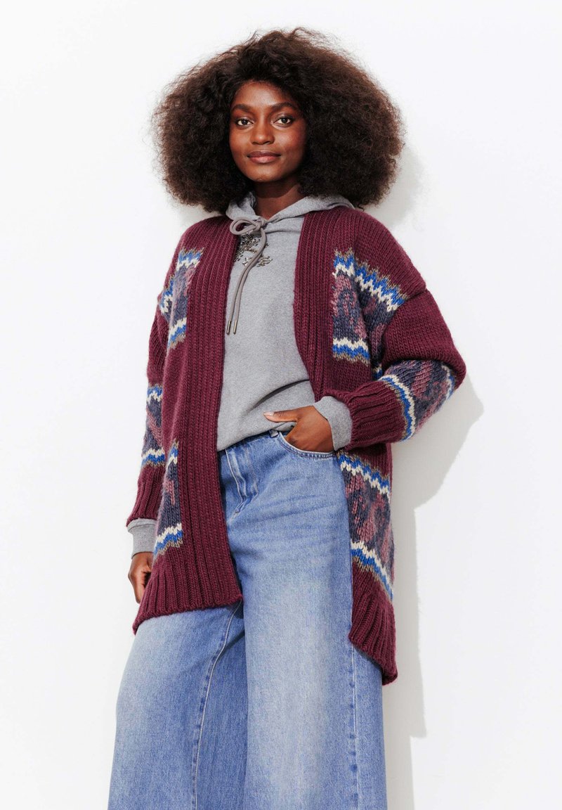 Cardigan en tricot bordeaux avec des motifs géométriques bleus et roses, porté par-dessus un sweat à capuche gris. Présente une coupe ample et des poignets côtelés.