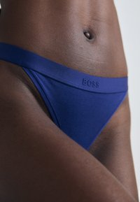 Close-up van een persoon die blauwe onderbroek draagt met "BOSS" op de tailleband, waarbij een deel van de torso en de bovenbenen zichtbaar is.