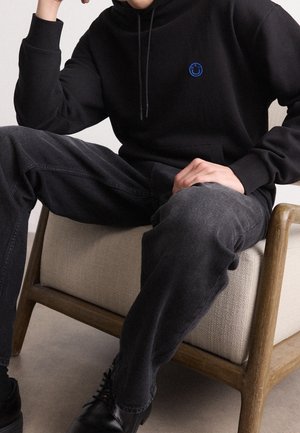Person siddende på beige polstret træstol iført sort hættetrøje med blåt logo, mørkegrå jeans og sorte lædersko.
