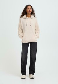 Světle béžová fleecová mikina s kapucí, stahovacími šňůrkami a předními kapsami, kombinovaná s černými kalhotami s širokými nohavicemi a béžovými sportovními botami.