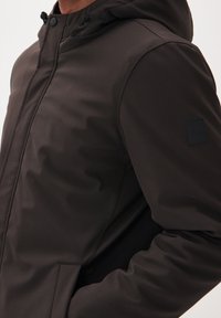 Mörkbrun huva jacka tillverkad av ett slätt, lätt material. Har tryckknappar och ett logomärke på överarmen.