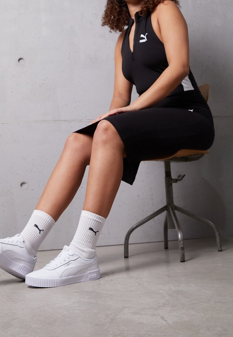 Robe noire sans manches avec fermeture éclair et accents blancs, associée à des baskets blanches et des chaussettes blanches côtelées. Fond en béton et chaise en métal.